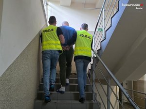 policjanci w odblaskowych kamizelkach z napisem Policja prowadzą po schodach zatrzymanego mężczyznę