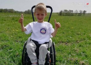 zdjęcie przedstawia 6-letniego chłopca siedzącego na wózku inwalidzkim i trzymającego kciuki do góry