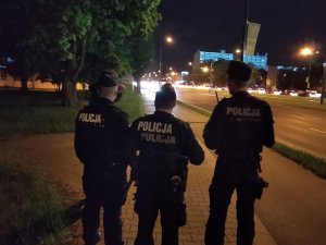 Na zdjęciu widocznych jest trzech policjantów ubranych w strój ćwiczebny nowego typu w czapkach typu furażerka. Policjanci pełnią służbę patrolową w okolicach stadionu miejskiego przy ul. Piłsudskiego w Łodzi. Zdjęcie wykonane w porze nocnej