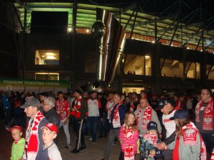 Na zdjęciu widoczna grupa kilkudziesięciu kibiców wychodząca ze stadionu po zakończeniu meczu. Kibice przemieszczają się obok pomnika z piłką znajdującego się przy stadionie. Zdjęcie wykonane zostało w porze nocnej
