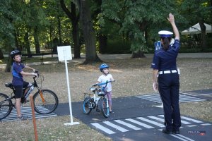 miasteczko ruchu drogowego, na pierwszym planie policjantka ruchu drogowego daje sygnał STOP, w tle dwoje dzieci z rowerami czeka na przejazd