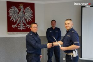 policjant otrzymuje podziękowanie od przełożonego, z tyłu godło Polski i funkcjonariusz w mundurze