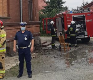 Policjant stoi obok budynku, z tyłu wóz straży pożarnej i strażacy