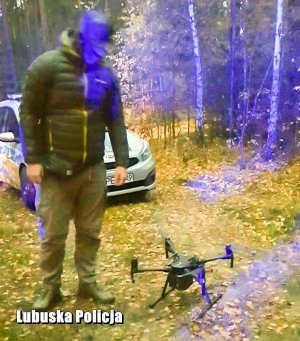 nieumundurowany funkcjonariusz stoi obok drona, za nim w tle radiowóz policyjny