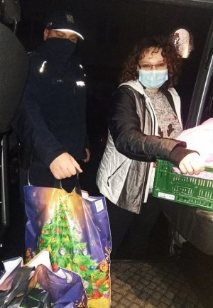 Po lewej stronie stoi umundurowany policjant, w ręku trzyma torbę ze świątecznym motywem. Po prawej stronie stoi kobieta, w rękach trzyma plastikowy kosz wypełniony przedmiotami