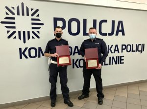 dwaj policjanci z podziękowaniami stoją na tle ściany z napisem Komenda Miejska Policji w Koszalinie