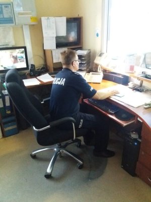policjant służby dyżurnej w koszulce z napisem Policja na plecach przy swoim stanowisku pracy. Przy nim stoi komputer i widoczne są telefony