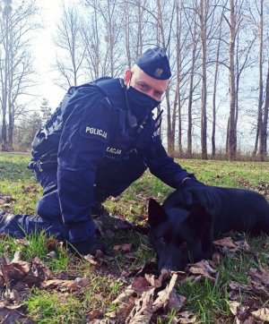 zdjęcie przedstawia policjanta z psem służbowym. Czarny owczarek niemiecki leży obok swojego przewodnika.