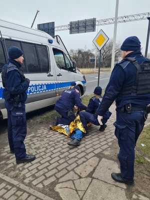 Policjanci udzielają pierwszej pomocy leżącemu na chodniku mężczyźnie. Obok widoczny jest policyjny furgon