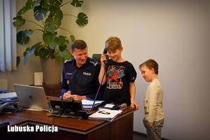 umundurowany funkcjonariusz pokazuje dzieciom swoje miejsce pracy, jeden z chłopców trzyma w ręku słuchawkę telefoniczną