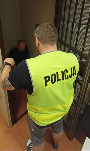 mężczyzna siedzi w celi a w drzwiach z kratami stoi policjant w żółtej kamizelce z napisem Policja na plecach