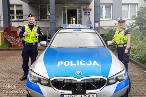 Dwoje umundurowanych policjantów stoi przy oznakowanym radiowozie