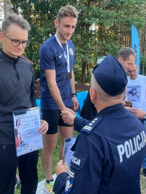 Po lewej stronie Arkadiusz Maruszewski na podium, stoi z dyplomem, stojący na wprost niego policjant w mundurze wręcza dyplom zwycięzcy na I miejscu