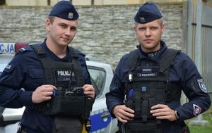 Dwóch policjantów stroi na tle radiowozu i budynku
