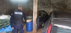W garażu stoi zaparkowany samochód, , leża jutowe worki obok stoi umundurowany policjant