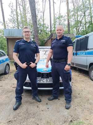 Na zdjęciu dwóch umundurowanych policjantów stoi na tle radiowozów