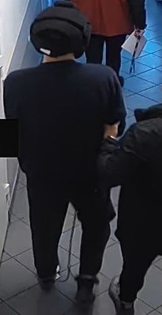 Na zdjęciu policjant prowadzi zatrzymanego mężczyznę. Zatrzymany na nogach ma kajdanki, a na głowie kask bezpieczeństwa