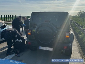 Stojący na autostradzie samochód marki Jeep oraz stojący przy nim funkcjonariusze Policji, w tym mundurowi z ruchu drogowego oraz siedzący na jezdni zatrzymany mężczyzna