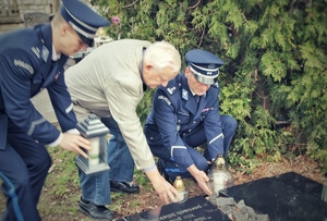na zdjęciu dwóch policjantów w umundurowaniu wyjściowym zapala znicze przy grobie, pomiędzy policjantami starszy mężczyzna również ze zniczem