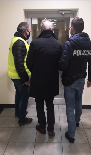 Policjanci w kamizelkach z napisem policja prowadzą mężczyznę, w tle przeszkolone drzwi