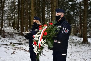 Złożenie kwiatów i zapalenie zniczy przy obelisku upamiętniającym katastrofę śmigłowca w Cisnej przez policjantów. Policjantka i policjant stoją przed obeliskiem z wieńcem i zniczami.