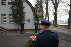 Na zdjęciu widoczny policjant stojący tyłem wraz z kwiatami, które zostaną złożone pod Tablicą Pamięci poświęconej podkomisarzowi Tadeuszowi Bartosik. Na wprost policjanta z kwiatami stoją dwaj policjanci, pomiędzy nimi znajduje się Tablica Pamięci.
