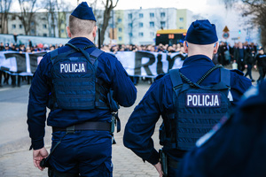 Umundurowani policjanci stojący tyłem, a przed nimi zebrani kibice