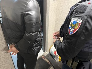 Na zdjęciu widać fragment sylwetki zatrzymanego mężczyzny w kajdankach, z prawej fragment sylwetki policjanta, a w tle drzwi wejściowe do pomieszczenia.