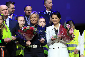 Policjantka i kobieta trzymają w ręku statuetkę oraz kwiaty. Za nimi inne osoby oraz dzieci w kamizelkach odblaskowych