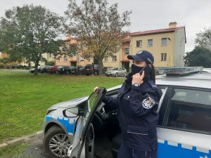 Umundurowana policjantka z maseczką na twarzy stoi przy radiowozie i trzyma przy uchu telefon komórkowy. W tle znajdują się dwa drzewa a za nimi budynek zaparkowane przy nim samochody osobowe.