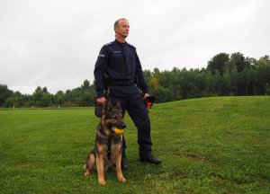 policjant stoi i patrzy w dal, obok siedzi przy jego nodze pies służbowy