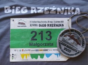 Na zdjęciu identyfikator biegu z nr startowym 213 i imieniem zawodniczki  Małgosia oraz medal po prawej stronie. U góry na zdjęciu widać napis Bieg Rzeźnika