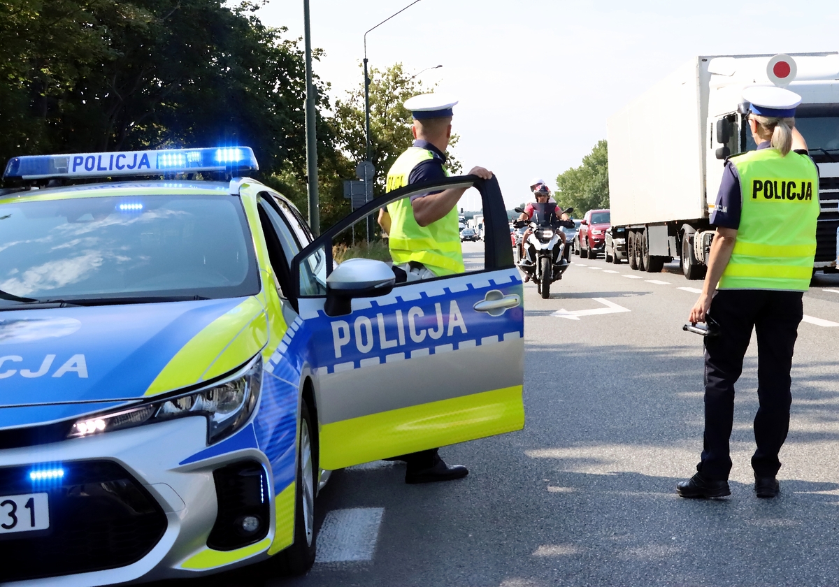 policjant i policjantka stoja przy radiowozie podczas kontroli drogowej. Po prawej stronie jadące drogą samochody. Do policjantów zbliża się mężczyzna jadący na motocykllu.