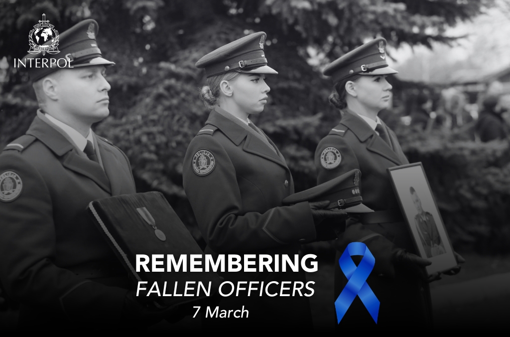 Zdjęcie czarno-białe. Dwie policjantki i policjant. Policjant z lewej strony trzyma pokrytą materiałem deskę, na którym znajduje się medal. Obok niego stoi policjantka trzymająca czapkę policyjną. Po prawej stronie policjantka trzymająca zdjęcie oprawione w ramę przedstawiające zmarłego policjanta. Poniżej napis: Remembering fallen officers 7 March. Po prawej stronie napisu niebieski kir.