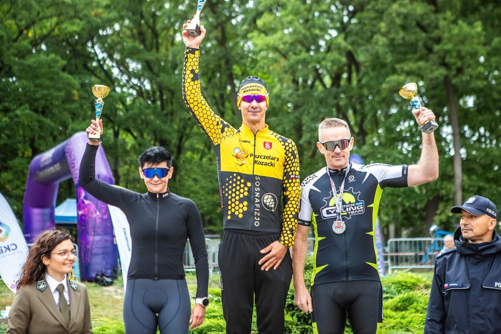 Zwycięzcy maratonu kolarskiego na podium trzymają w rękach puchary.