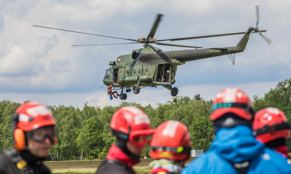 Mi-8 unosi się nad głowami strażaków, u jego burty zwisa na linie jeden z ich kolegów.