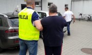 Zatrzymany mężczyzna tyłem w czarnej koszulce i czarnych dresowych spodniach z czerwonymi lampasami odprowadzany przez policjanta przez parking przed komendą