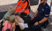 policjant z ratownikiem przy fantomie pokazują dzieciom jak udzielać pierwszej pomocy