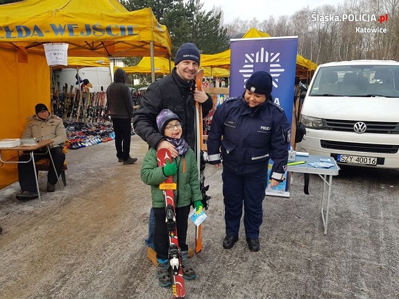 stoi chłopczyk trzymający narty oraz ulotkę akcji &quot;Kręci mnie bezpieczeństwo na stoku&quot; wraz z nim stoi ojciec również trzymający narty obok stoi policjantka