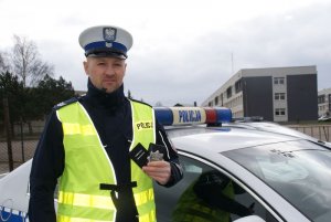 Policjant przy radiowozie trzyma legitymację policyjną i legitymację egzaminatora