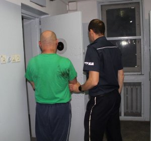 kolorowe zdjęcie przedstawiające umundurowanego policjanta prowadzącego do celi  PDOZ zatrzymanego do sprawy posiadania środków odurzających