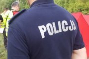 policjant stoi tyłem, na koszulce na plecach widnieje napis policja, w tle widać technika kryminalistyki w kamizelce odblaskowej, widać również czerwony parawan