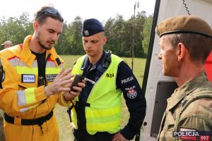 Na zdjęciu ratownik z grupy ratowniczej „Nadzieja” w żółtym kombinezonie udziela Policjantowi i żołnierzowi WOT instrukcji używania narzędzia do nawigacji GPS .