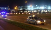 Na zdjęciu wykonanym w porze nocnej, widoczne są dwa oznakowane radiowozy policji, które jadą na pasie ruchu w okolicach stadionu miejskiego przy ul. Piłsudskiego w Łodzi. W tle widać rozświetlony stadion w trakcie trwania meczu