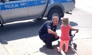 policjant rozmawia z małą dziewczynką, która prowadzi wózek, w tle stoi radiowóz