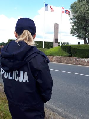 Umundurowana polska policjantka. W tle widoczny pomnik oraz powiewające flagi Francji i Norwegii