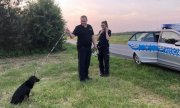 policjant i policjantka stoją przy radiowozie z psem