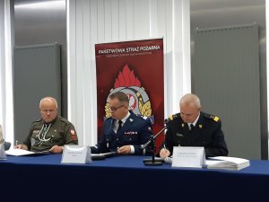 Umundurowani Pierwszy Zastępca Komendanta Głównego, Komendant Główny Państwowej Straży Pożarnej oraz przedstawiciel harcerzy podpisują Protokół