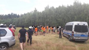 policjanci z OPP w Łodzi oraz otoczyli grupa pseudokibiców ubranych w pomarańczowe koszulki, po prawej stronie radiowóz policyjny a w głębi las