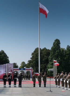 Na pierwszym planie maszt z powiewającą flagą narodową, obok stojący żołnierze, w tle ododdziały zmierzają w kierunku Grobu Nienznego Żołnierza